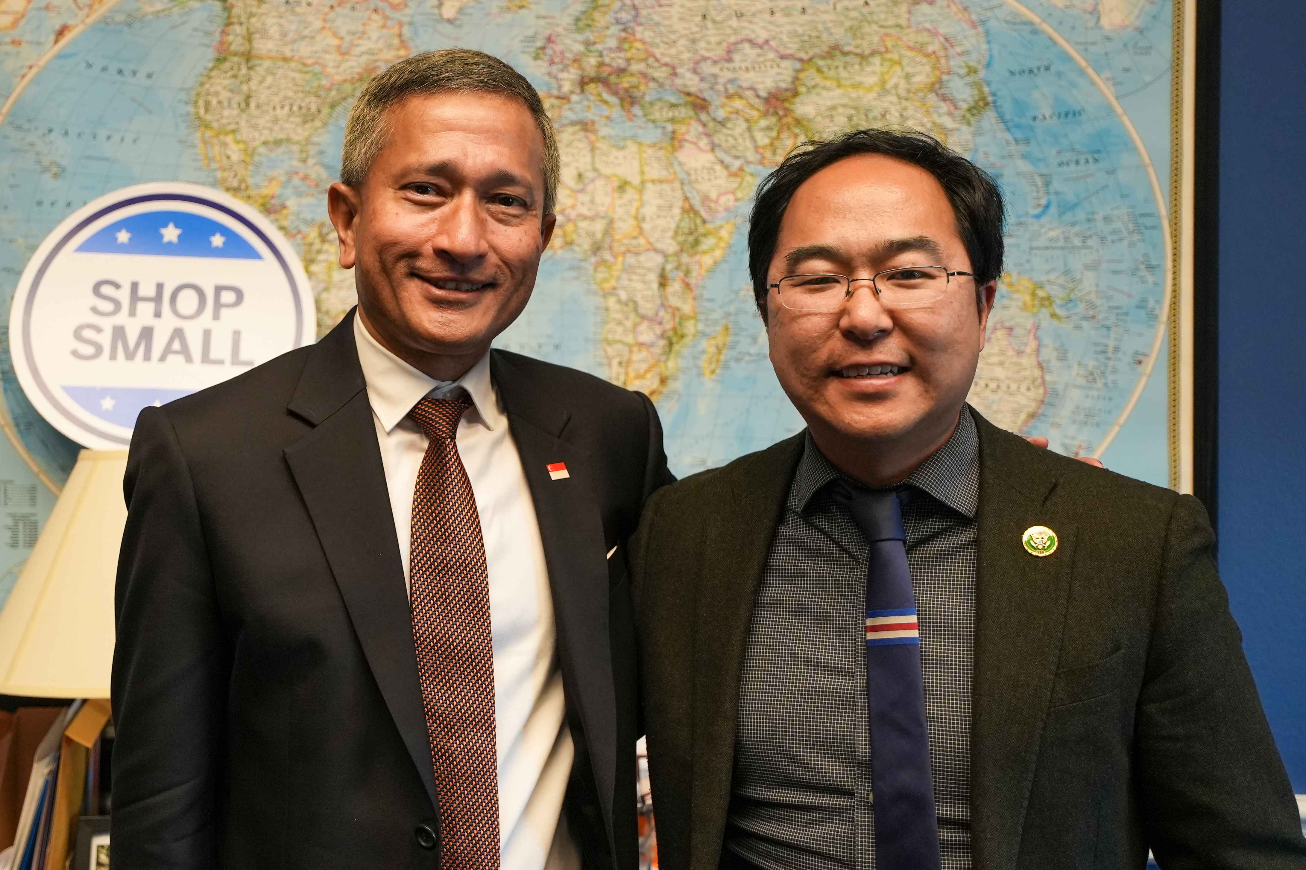 Two formally dressed men with respective country pins stand in front of a world map.